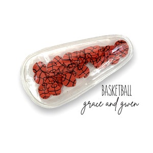 May include: Clear plastic hair clip filled with red and black basketball-themed confetti. The text "BASKETBALL grace and guen" is printed below the clip.