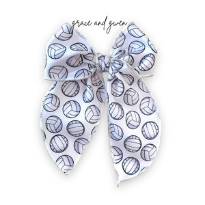 May include: White bow with a silver glitter edge and a pattern of black and white volleyballs. The bow is tied in a large loop with the ends of the bow hanging down.