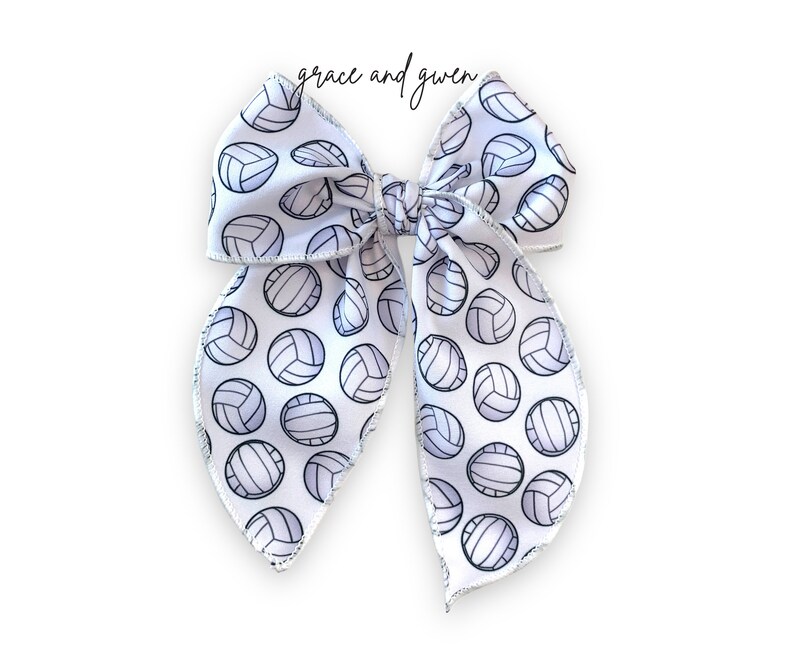 May include: White bow with a silver glitter edge and a pattern of black and white volleyballs. The bow is tied in a large loop with the ends of the bow hanging down.