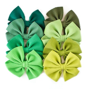 May include: An assortment of eight green fabric bows in various shades, from deep emerald to light lime. The bows are arranged closely together on a white background, showcasing their textured surface and symmetrical design. These accessories are ideal for hair styling or crafting.
