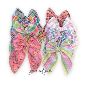 May include: A set of eight pastel-colored hair bows with various Easter-themed patterns, including plaid, stripes, and bunny designs. The bows are arranged in a stack, with the text "grace and gwen" visible at the bottom.