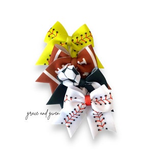 May include: Three hair bows with different sports themes. The top bow is yellow with a softball design. The middle bow is brown with a football and soccer ball design. The bottom bow is white with a baseball design.