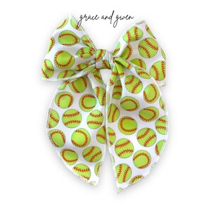 May include: A white fabric bow with a green and red softball pattern. The bow is tied in a knot and has a loop on the top.