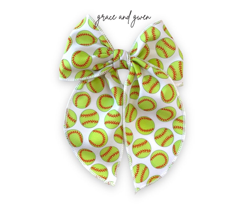 May include: A white fabric bow with a green and red softball pattern. The bow is tied in a knot and has a loop on the top.