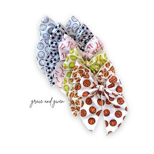May include: A set of five hair bows with different sports ball prints. The bows are made of white satin with a variety of sports balls printed on them, including basketball, baseball, softball, and volleyball. The bows are tied together with a white ribbon.