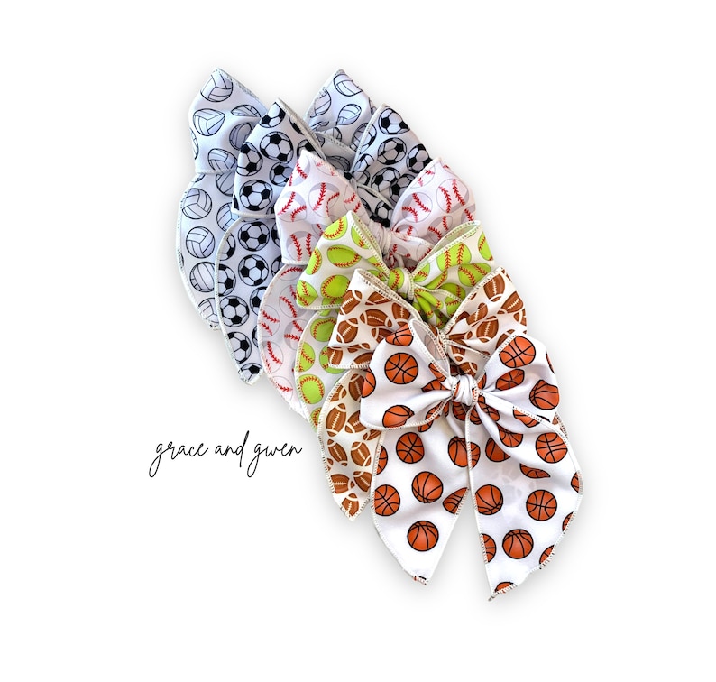 May include: A set of five hair bows with different sports ball prints. The bows are made of white satin with a variety of sports balls printed on them, including basketball, baseball, softball, and volleyball. The bows are tied together with a white ribbon.