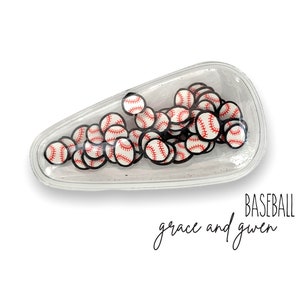 May include: A clear plastic hair clip filled with small baseball-themed charms. The clip is shaped like a teardrop and has a white background. The text "BASEBALL grace and gwen" is printed below the clip.