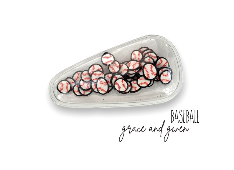 May include: A clear plastic hair clip filled with small baseball-themed charms. The clip is shaped like a teardrop and has a white background. The text "BASEBALL grace and gwen" is printed below the clip.