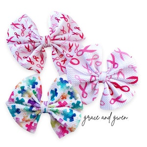 May include: Three hair bows with different patterns. One bow has a pink ribbon pattern, another has a pink ribbon pattern with a small scissor design, and the last bow has a colorful puzzle piece pattern. The bows are on a white background with the text "grace and gwen" in black.