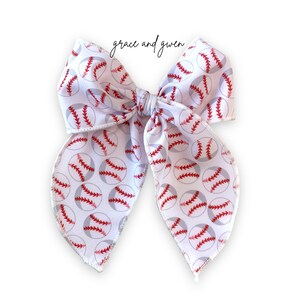 May include: White bow with a baseball print. The bow is tied in a large loop with two tails. The baseballs are red and gray with white stitching.