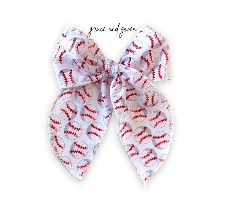May include: White bow with a baseball print. The bow is tied in a large loop with two tails. The baseballs are red and gray with white stitching.