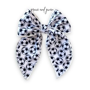 May include: A white hair bow with a black and white soccer ball pattern. The bow has a white satin trim.