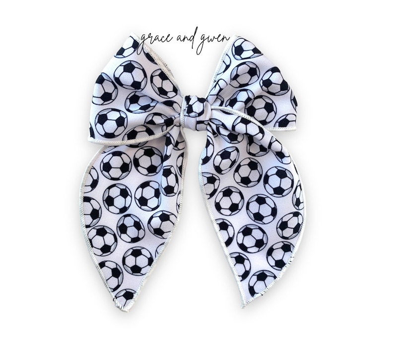 May include: A white hair bow with a black and white soccer ball pattern. The bow has a white satin trim.