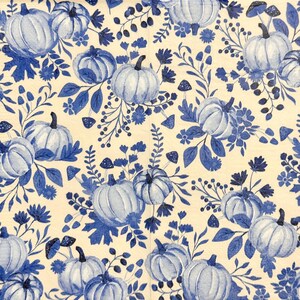 May include: A repeating pattern of blue pumpkins, flowers, and foliage on a cream background. The pumpkins have dark stems and are surrounded by blue leaves, flowers, and mushrooms. The design is reminiscent of a watercolor painting.