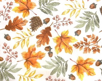 May include: A white paper napkin featuring a repeating pattern of autumn elements including orange and yellow maple leaves, brown oak leaves, green fern-like foliage, small brown acorns, and dark brown pinecones scattered across the surface.