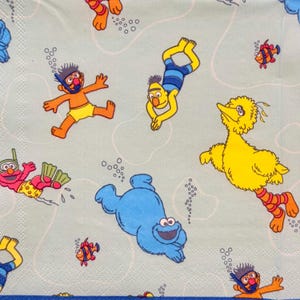 May include: A light blue paper napkin featuring cartoon characters from Sesame Street. The design includes Ernie, Bert, Elmo, Cookie Monster, and Big Bird, all depicted as if swimming underwater, surrounded by bubbles and fish.