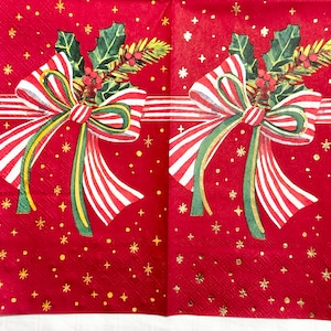 May include: Two red Christmas napkins with a festive design. Each napkin features a large bow with red and white stripes, green and gold accents, and holly and berry sprigs. Gold stars and dots are scattered across the red background.