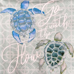 May include: A light blue napkin featuring two watercolor sea turtles. The turtles are in shades of blue and gray, with the words "Go with the Flow" in white script. The background has a subtle geometric pattern.