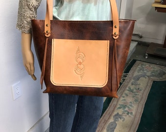 May include: A brown leather tote bag with a light brown leather pocket on the front. The pocket has a decorative design in the center. The bag has a long strap and a zipper closure.