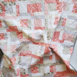 以下が含まれることがあります： パッチワークデザインの手作りキルト。ピンク、白、グレーの色合いの正方形が特徴で、花柄と幾何学模様があります。ボーダーは白と黒のストライプです。キルトは柔らかい生地で作られています。