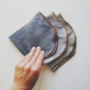 May include: A stack of four square, soft, baby washcloths in shades of blue and green. Each cloth has a brown stitched border. The washcloths are arranged in a fan shape, with a hand gently touching the top cloth.