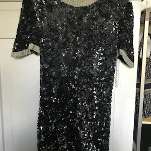 Könnte beinhalten: Ein schwarzes Pailletten-Minikleid mit kurzen Ärmeln und einem silbernen Besatz an Ausschnitt und Ärmeln.