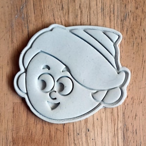 May include: White plastic cookie cutter shaped like a cartoon character with a party hat. The character has large eyes, a smile, and a single curl of hair.