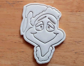 May include: White plastic cookie cutter shaped like the Grinch from the movie How the Grinch Stole Christmas. The Grinch is smiling with his eyes closed.