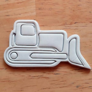 May include: White plastic cookie cutter shaped like a bulldozer. The cutter has a detailed outline of the bulldozer, including the cab, tracks, and blade.