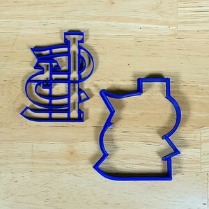 K&ouml;nnte beinhalten: Zwei blaue Plastik-Ausstecher, einer in Form des Buchstabens "STL" und der andere in Form einer Baseballkappe.