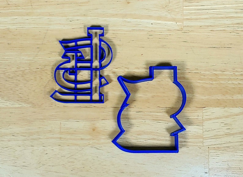 K&ouml;nnte beinhalten: Zwei blaue Plastik-Ausstecher, einer in Form des Buchstabens "STL" und der andere in Form einer Baseballkappe.