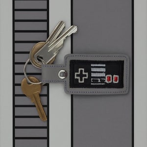 May include: A gray leather keychain with a black embroidered video game controller design. The keychain is attached to a set of silver and gold keys. The controller design features a plus sign, lines, and buttons.
