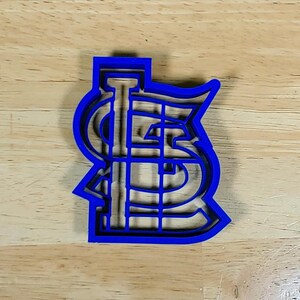 K&ouml;nnte beinhalten: Ein blauer Plastik-Ausstecher in Form des Logos der St. Louis Cardinals. Das Logo ist ein stilisiertes "STL" mit einem Baseballschl&auml;ger in der Mitte.