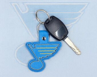 Pode incluir: Um porta-chaves bordado azul e amarelo com uma chave de carro. O porta-chaves apresenta um emblema estilizado com fundo azul e detalhes amarelos. A chave do carro é preta e prateada, presa ao porta-chaves com um anel de metal.