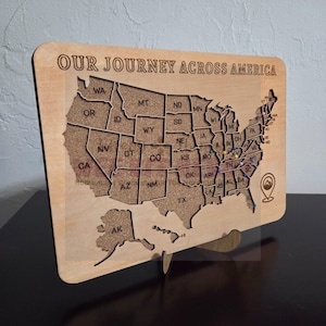 May include: Wooden map of the United States with state outlines and cork interior. The text "OUR JOURNEY ACROSS AMERICA" is engraved at the top. Includes a stand for display and colorful pins marking locations.