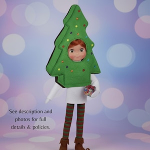 Op de afbeelding: Een pop verkleed als groene kerstboom met kleurrijke geborduurde versieringen. De pop heeft rood haar en houdt een klein cadeautje vast. Ze draagt een wit shirt, gestreepte leggings en bruine laarzen. De achtergrond is zachtblauw met witte bokeh.