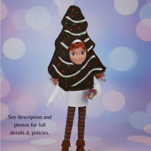 May include: A doll dressed in a brown Christmas tree costume with white trim and red and green accents. The doll is wearing a white dress, striped green and red tights, and brown boots. The doll is holding a small gift. The text in the image says "See description and photos for full details & policies."