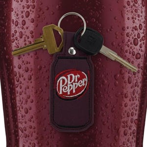 May include: A burgundy leather key fob with the Dr. Pepper logo in red and white embroidery. Two keys are attached to the silver keyring. The background is a textured, dark red surface.