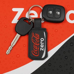 May include: A black car key, key fob, and a Coca-Cola Zero keychain on a red and white background. The keychain is black with the Coca-Cola Zero logo in red and white. The key fob has two buttons.