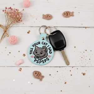 May include: A light blue cat-themed keychain with the text "TELL YOUR CAT I SAD PSPSPS". The keychain has a silver ring and is attached to a black car key. Decorative elements include pink yarn balls and small fish-shaped ornaments.