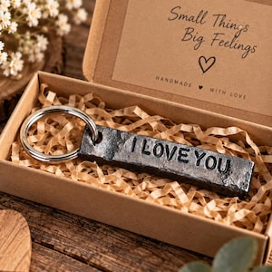 May include: A rectangular dark gray metal keychain with the stamped text I LOVE YOU attached to a silver circular ring. The keychain is presented inside a brown cardboard gift box filled with shredded paper, featuring the text Small Things Big Feelings.