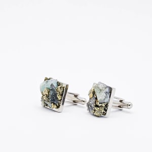 May include: A pair of silver cufflinks with a square design. Each cufflink features a cluster of green and grey stones with gold accents.