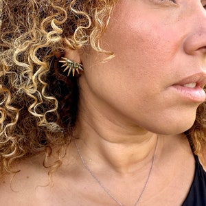 May include: A gold necklace with a starburst pendant and matching earrings. The necklace is on a person with curly blonde hair.