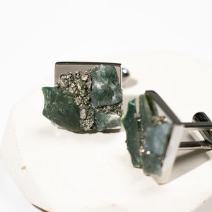May include: A pair of silver cufflinks with green and silver stone inlays. The stones are irregularly shaped and appear to be a mix of green agate and pyrite.