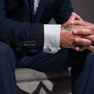 May include: A man wearing a dark blue suit with a white shirt and cufflinks. He has a tattoo of a flower on his wrist.