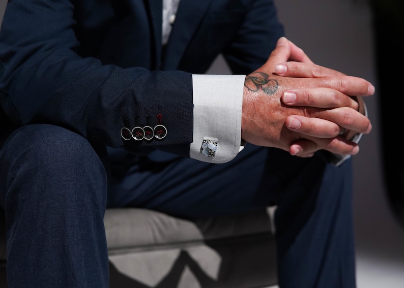 May include: A man wearing a dark blue suit with a white shirt and cufflinks. He has a tattoo of a flower on his wrist.