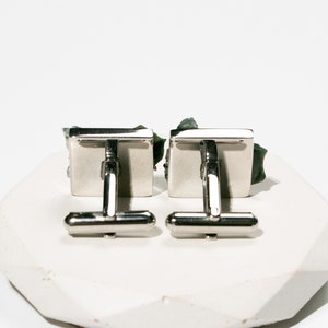 May include: A pair of silver cufflinks with green stone accents. The cufflinks are square shaped and have a polished finish.