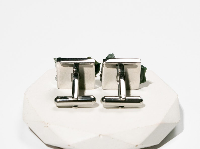 May include: A pair of silver cufflinks with green stone accents. The cufflinks are square shaped and have a polished finish.