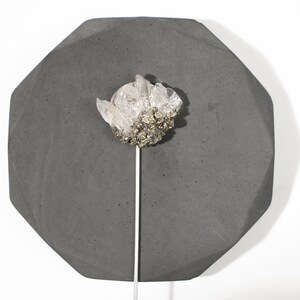 May include: A cluster of clear quartz crystals with a base of pyrite, mounted on a silver rod, displayed on a dark gray octagonal concrete base.