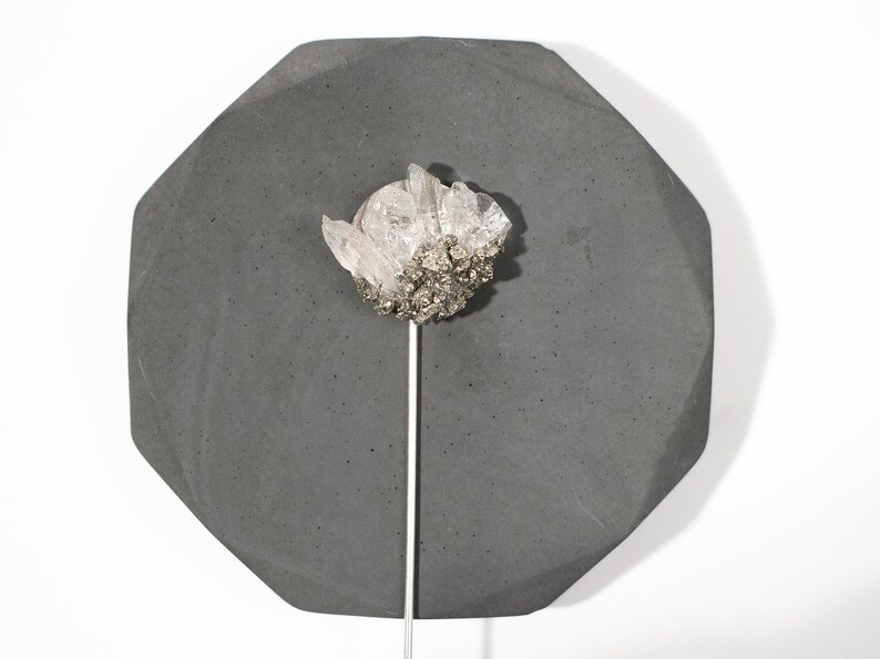 May include: A cluster of clear quartz crystals with a base of pyrite, mounted on a silver rod, displayed on a dark gray octagonal concrete base.